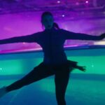 A skater does a pirouette while the scene around her is a dazzling mix of purple, green, and blue light