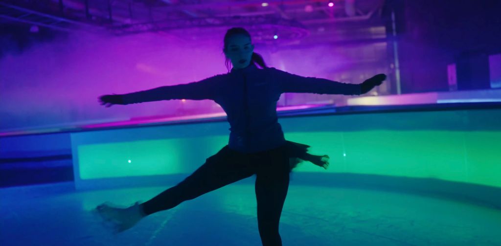 A skater does a pirouette while the scene around her is a dazzling mix of purple, green, and blue light