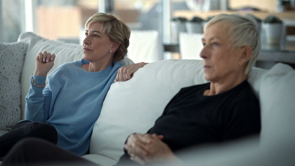 Chris Everts and Martina Navratilova sit on a couch. They are watching a TV that is outside the frame and have animated expressions on their faces. They are both white women in their late 60s with short-cropped blond hair. The couch is white and the room is bright.