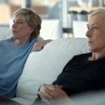 Chris Everts and Martina Navratilova sit on a couch. They are watching a TV that is outside the frame and have animated expressions on their faces. They are both white women in their late 60s with short-cropped blond hair. The couch is white and the room is bright.