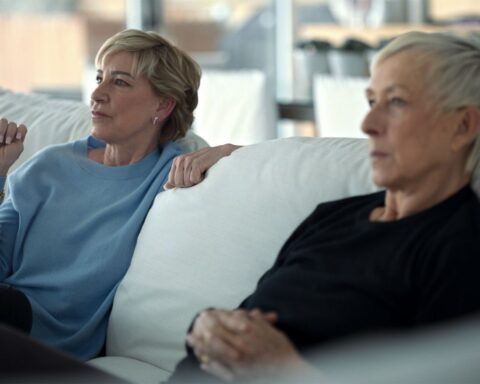 Chris Everts and Martina Navratilova sit on a couch. They are watching a TV that is outside the frame and have animated expressions on their faces. They are both white women in their late 60s with short-cropped blond hair. The couch is white and the room is bright.
