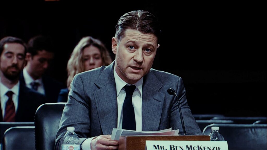 Actor Ben McKenzie sits at a table testifying in a hearing. He is a white man in his 40s with brown hair, wearing a grey suit and dark tie. He is looking toward the camera and reading a statement