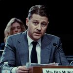 Actor Ben McKenzie sits at a table testifying in a hearing. He is a white man in his 40s with brown hair, wearing a grey suit and dark tie. He is looking toward the camera and reading a statement