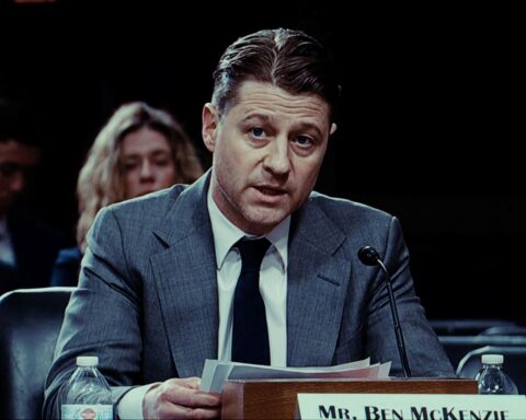 Actor Ben McKenzie sits at a table testifying in a hearing. He is a white man in his 40s with brown hair, wearing a grey suit and dark tie. He is looking toward the camera and reading a statement