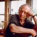 Filmmaker Frederick Wiseman is pictured sitting at a desk with one hand raised to prop up his head and the other arm resting on the desk. He is a white man in his 80s with curly grey hair, and is wearing a black golf shirt. He is looking directly into the camera. The background shows a bright, rustic room with big windows.