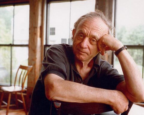 Filmmaker Frederick Wiseman is pictured sitting at a desk with one hand raised to prop up his head and the other arm resting on the desk. He is a white man in his 80s with curly grey hair, and is wearing a black golf shirt. He is looking directly into the camera. The background shows a bright, rustic room with big windows.
