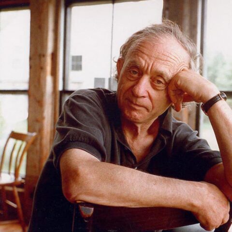 Filmmaker Frederick Wiseman is pictured sitting at a desk with one hand raised to prop up his head and the other arm resting on the desk. He is a white man in his 80s with curly grey hair, and is wearing a black golf shirt. He is looking directly into the camera. The background shows a bright, rustic room with big windows.