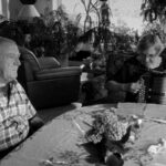 A black and white photo of an older man and woman sit at a dining room table. She is playing the according and looking down, while he sits listening with his eyes closed.
