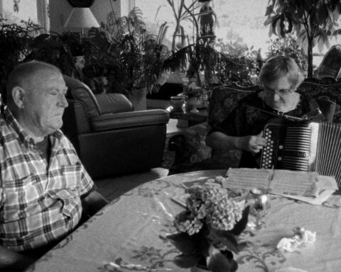 A black and white photo of an older man and woman sit at a dining room table. She is playing the according and looking down, while he sits listening with his eyes closed.
