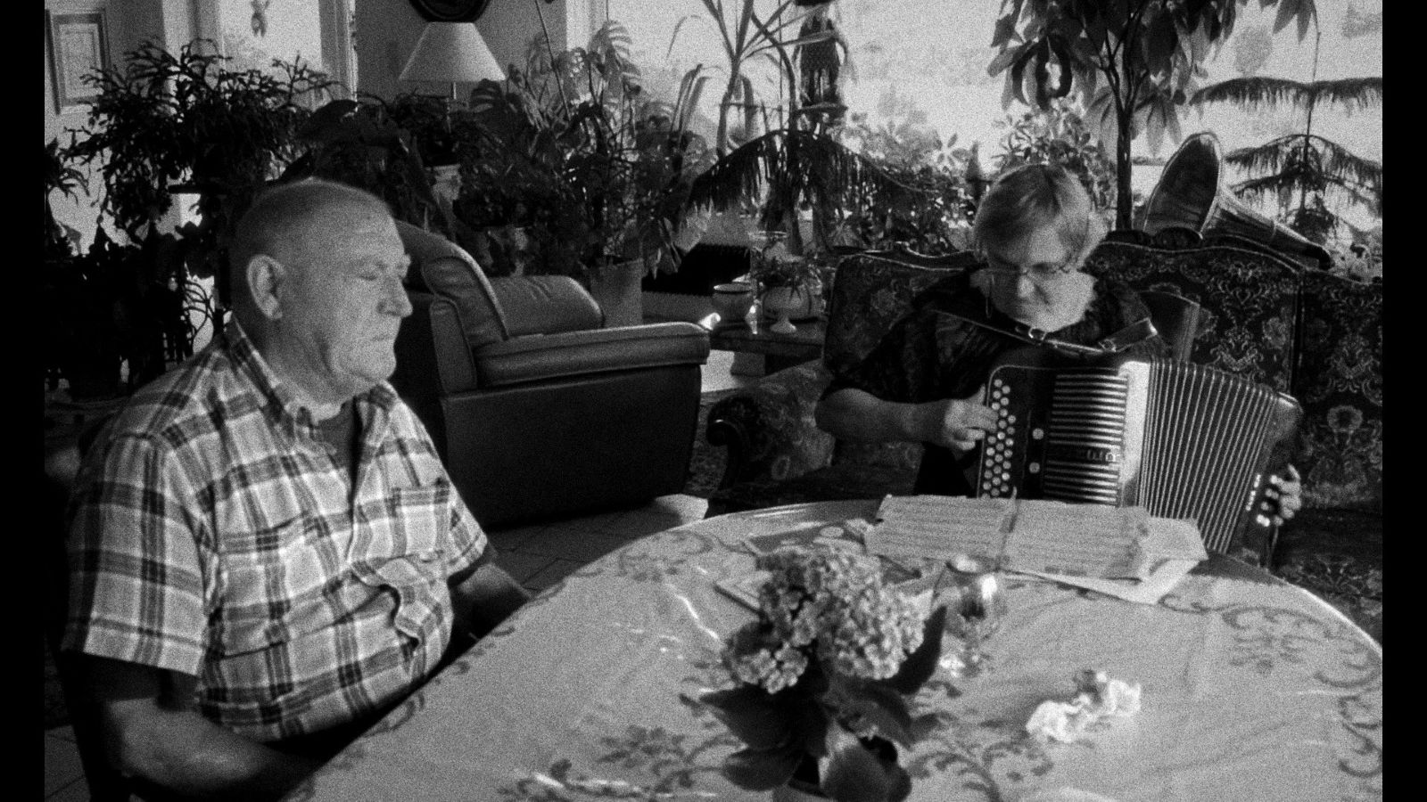 A black and white photo of an older man and woman sit at a dining room table. She is playing the according and looking down, while he sits listening with his eyes closed.