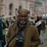 Photographer Misan Harriman is pictured standing in front of a group of protesters. He is a Black man with a grey beard and moustache. He is wearing a green jacket and has a camera hanging over his chest. The protesters behind him are out of focus.