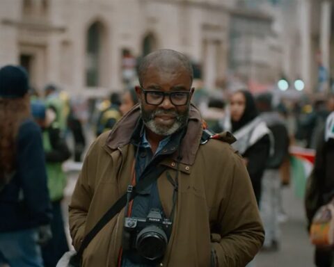 Photographer Misan Harriman is pictured standing in front of a group of protesters. He is a Black man with a grey beard and moustache. He is wearing a green jacket and has a camera hanging over his chest. The protesters behind him are out of focus.