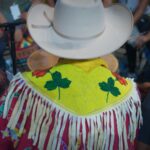 A Blackfoot man is pictured from behind as we see him dressed for a rodeo. He is wearing a white cowboy hat and a yellow beaded shawl with two green leaves and two red ones among the bead work. The garmet has white fringe that hangs down from his shoulders. He is wearing a red shirt underneath.