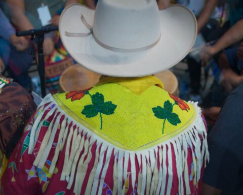 A Blackfoot man is pictured from behind as we see him dressed for a rodeo. He is wearing a white cowboy hat and a yellow beaded shawl with two green leaves and two red ones among the bead work. The garmet has white fringe that hangs down from his shoulders. He is wearing a red shirt underneath.