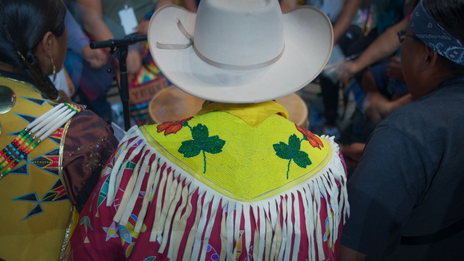 A Blackfoot man is pictured from behind as we see him dressed for a rodeo. He is wearing a white cowboy hat and a yellow beaded shawl with two green leaves and two red ones among the bead work. The garmet has white fringe that hangs down from his shoulders. He is wearing a red shirt underneath.