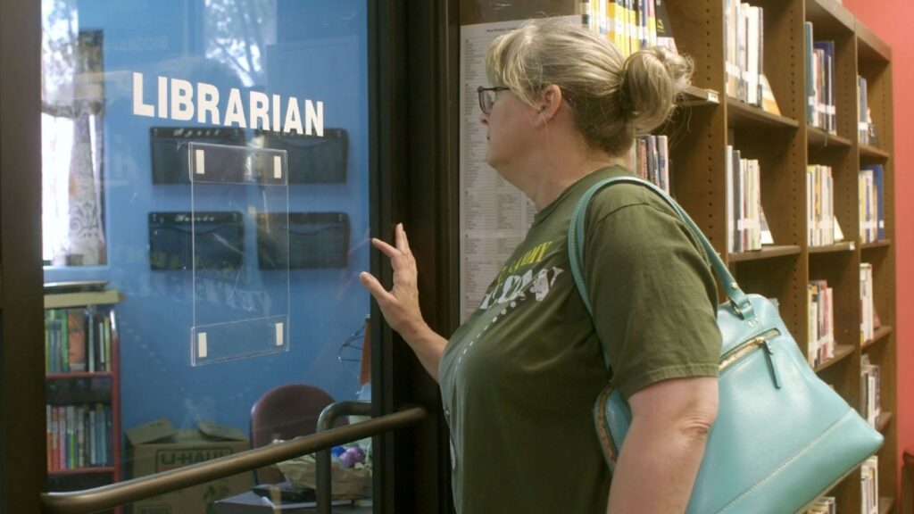A white woman with blond hair and a green t-shirt stands in a library. She is facing the window of an empty office with the word 'librarian' written in white letters on the glass. There are many shelves of books in the background.