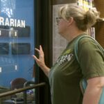 A white woman with blond hair and a green t-shirt stands in a library. She is facing the window of an empty office with the word 'librarian' written in white letters on the glass. There are many shelves of books in the background.
