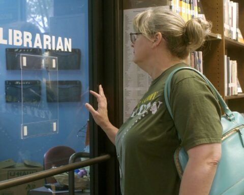A white woman with blond hair and a green t-shirt stands in a library. She is facing the window of an empty office with the word 'librarian' written in white letters on the glass. There are many shelves of books in the background.