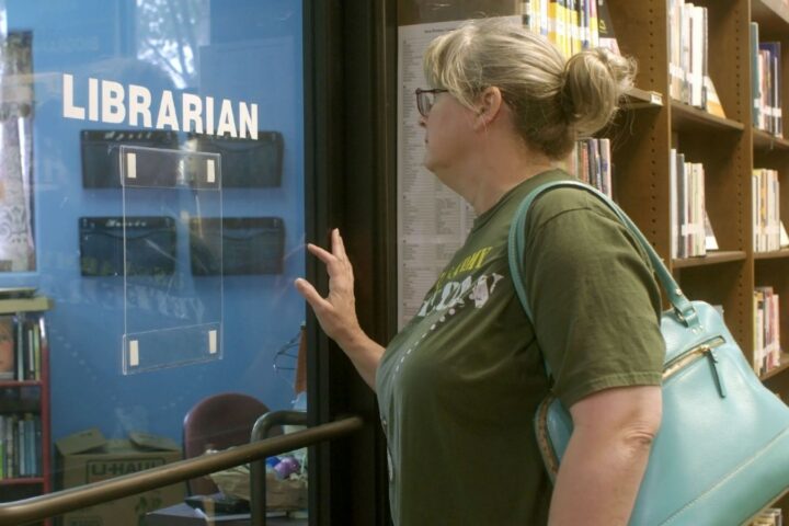 A white woman with blond hair and a green t-shirt stands in a library. She is facing the window of an empty office with the word 'librarian' written in white letters on the glass. There are many shelves of books in the background.