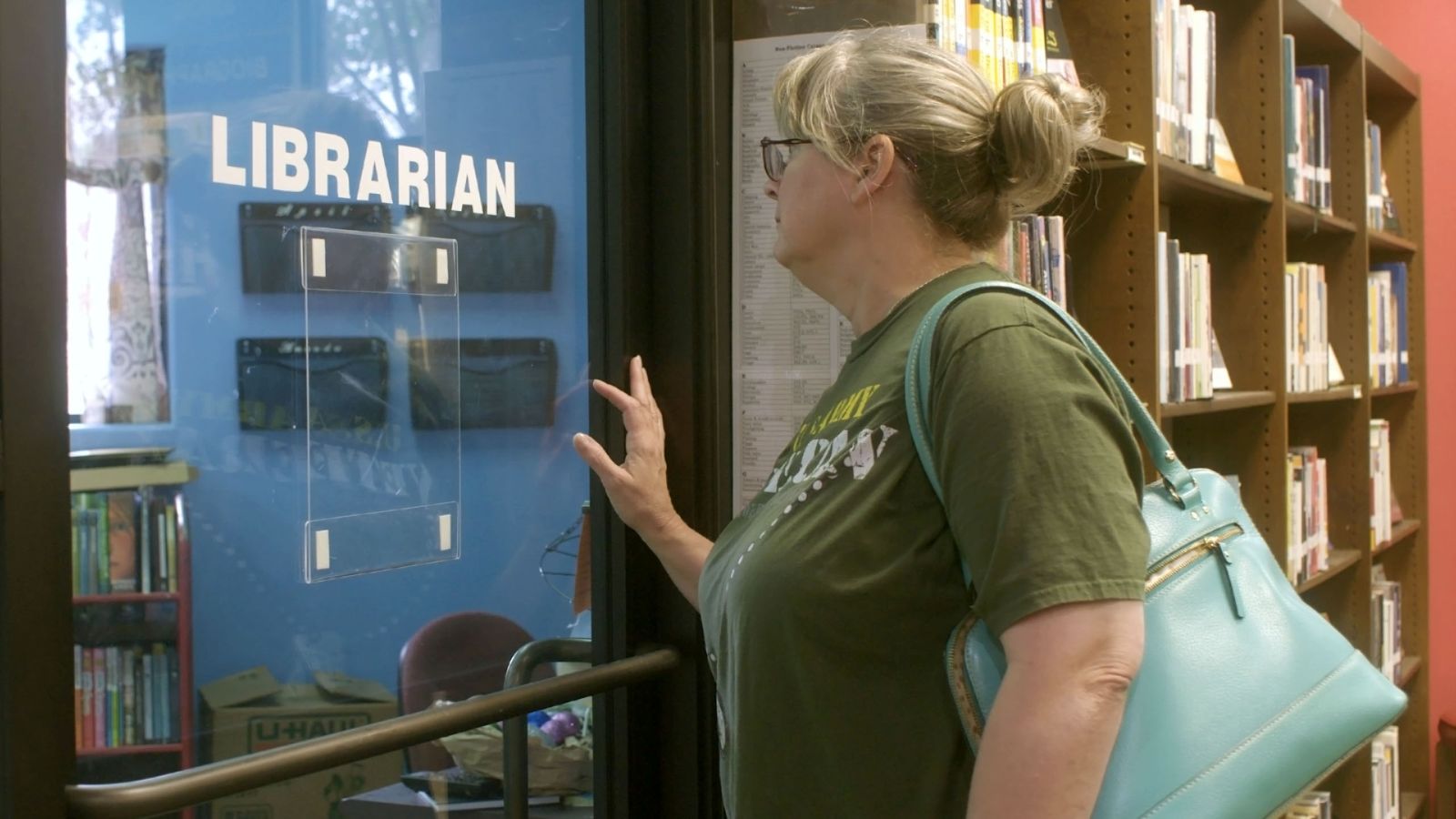 A white woman with blond hair and a green t-shirt stands in a library. She is facing the window of an empty office with the word 'librarian' written in white letters on the glass. There are many shelves of books in the background.