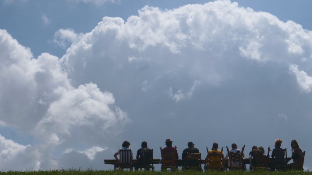 A wide shot shows a many people gathered around a larger dinner table atop a New Orleans levee. They are seen in silhouette against a backdrop of an approaching storm cloud on a sunny day.