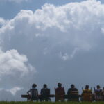 A wide shot shows a many people gathered around a larger dinner table atop a New Orleans levee. They are seen in silhouette against a backdrop of an approaching storm cloud on a sunny day.
