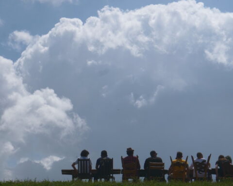 A wide shot shows a many people gathered around a larger dinner table atop a New Orleans levee. They are seen in silhouette against a backdrop of an approaching storm cloud on a sunny day.