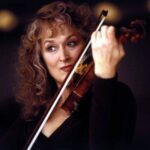 Meryl Streep is pictured in close-up. She is playing the violin and has an intense facial expression. She is wearing a black dress and is photographed against a black background. She is a white woman with curly auburn hair.