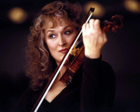 Meryl Streep is pictured in close-up. She is playing the violin and has an intense facial expression. She is wearing a black dress and is photographed against a black background. She is a white woman with curly auburn hair.