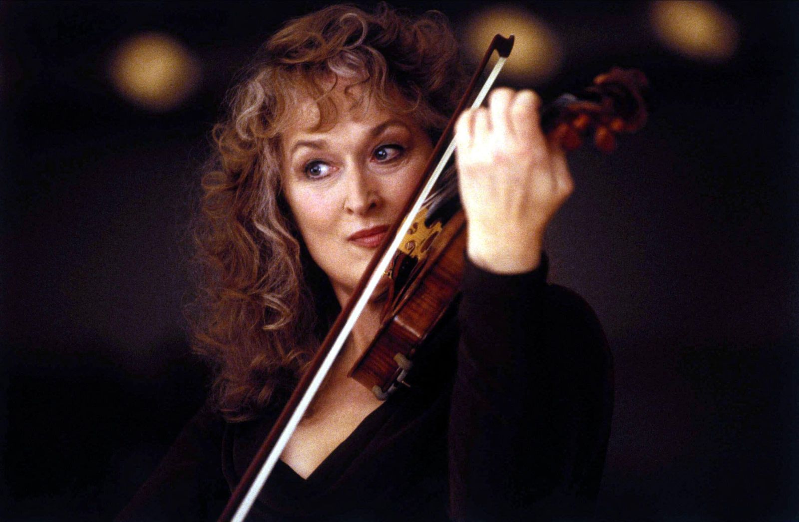 Meryl Streep is pictured in close-up. She is playing the violin and has an intense facial expression. She is wearing a black dress and is photographed against a black background. She is a white woman with curly auburn hair.