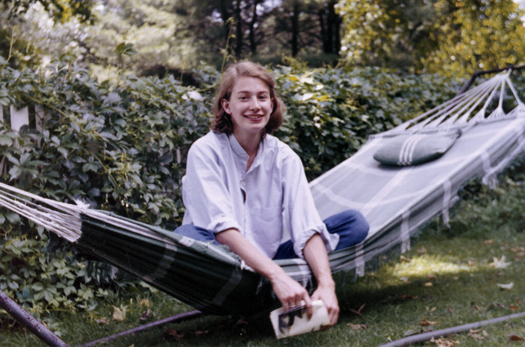 An archival photo shows poet Mary Oliver sitting cross-legged on a hammock. She is a white woman with brown hair, wearing a white collared shirt and blue jeans. She is holding a book and there is a hedge behind her.