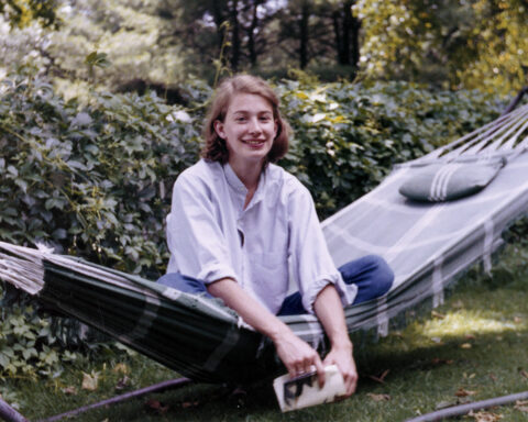 An archival photo shows poet Mary Oliver sitting cross-legged on a hammock. She is a white woman with brown hair, wearing a white collared shirt and blue jeans. She is holding a book and there is a hedge behind her.