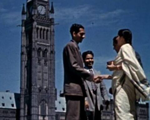 An archival image shows a group of people standing in front of Parliament Hill in Ottawa. The shot is from a low angle as it observes the people talking and shaking hands as the Peace Tower looms behind them.