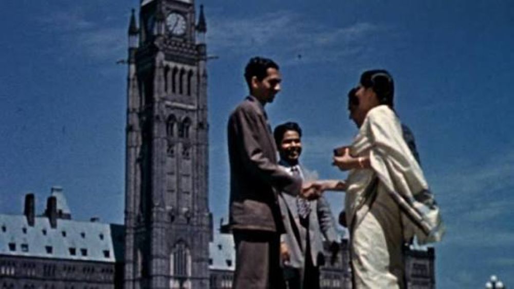 An archival image shows a group of people standing in front of Parliament Hill in Ottawa. The shot is from a low angle as it observes the people talking and shaking hands as the Peace Tower looms behind them.