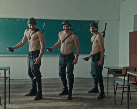 Three soldiers pose in a classroom as they hold grenades and face the camera. They are white men without shirts and are wearing green army pants, helmets, and have rifles strapped to their backs. They are standing in a line and there is a chalkboard behind them.