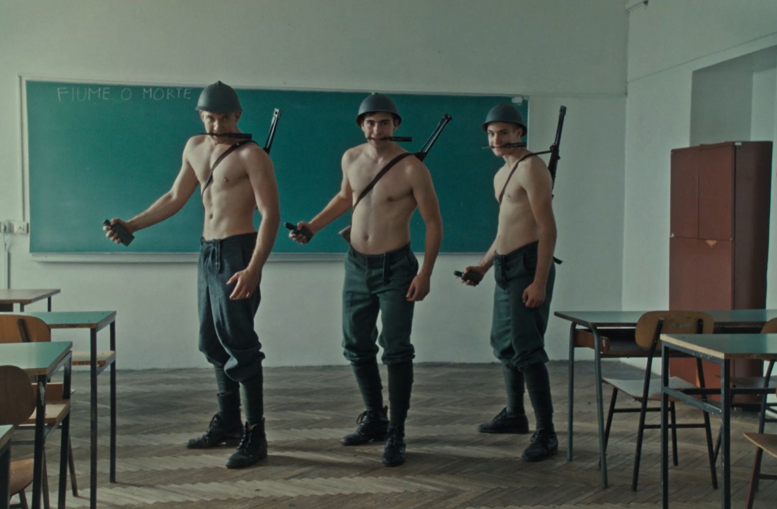 Three soldiers pose in a classroom as they hold grenades and face the camera. They are white men without shirts and are wearing green army pants, helmets, and have rifles strapped to their backs. They are standing in a line and there is a chalkboard behind them.