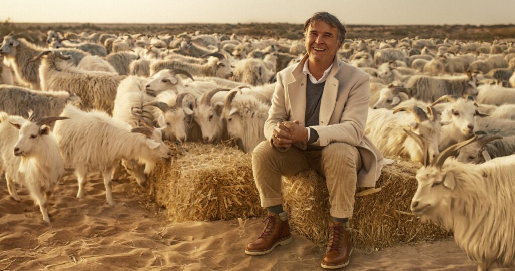 Fashion mogul Brunello Cucinelli is an Italian man in his early 70s. He is pictured sitting on a bale of hay surrounded by a herd of goats. He is wearing a beige jacket and grey sweater with tan pants. He has grey hair and is smiling.