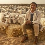 Fashion mogul Brunello Cucinelli is an Italian man in his early 70s. He is pictured sitting on a bale of hay surrounded by a herd of goats. He is wearing a beige jacket and grey sweater with tan pants. He has grey hair and is smiling.