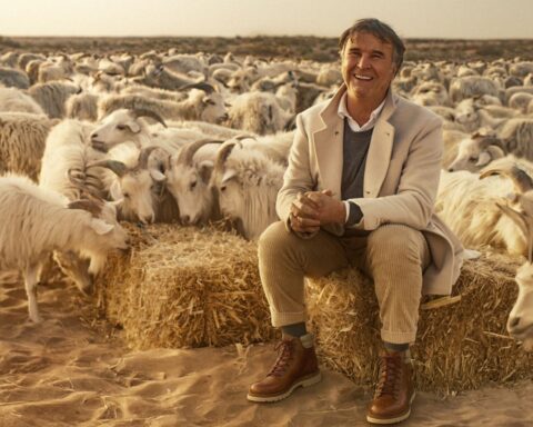 Fashion mogul Brunello Cucinelli is an Italian man in his early 70s. He is pictured sitting on a bale of hay surrounded by a herd of goats. He is wearing a beige jacket and grey sweater with tan pants. He has grey hair and is smiling.