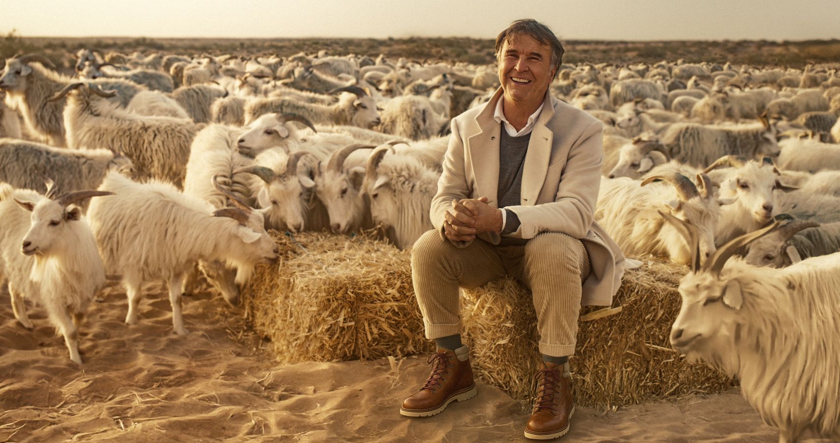Fashion mogul Brunello Cucinelli is an Italian man in his early 70s. He is pictured sitting on a bale of hay surrounded by a herd of goats. He is wearing a beige jacket and grey sweater with tan pants. He has grey hair and is smiling.