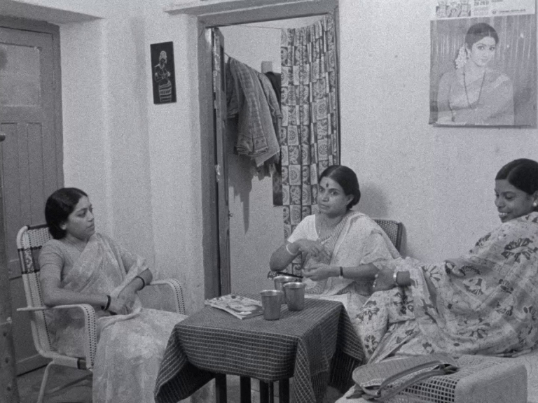A wide black and white shot shows three women sitting in a living room of a small, modest home. They are having a conversation.