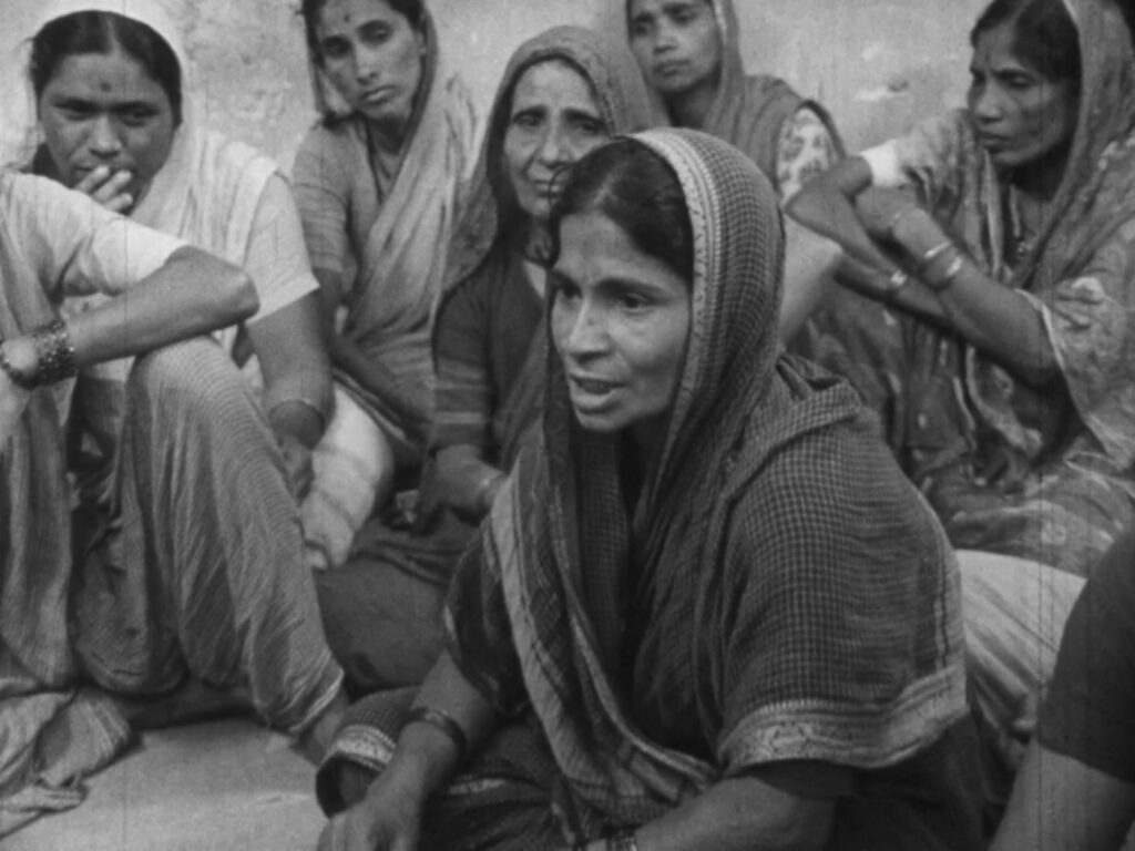 A black and white image shows an Indian woman seated and speaking to a group of women who are listening to her. She is wearing a dark robe and her head is covered.
