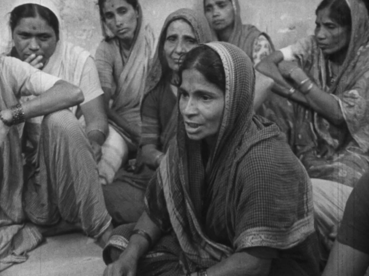 A black and white image shows an Indian woman seated and speaking to a group of women who are listening to her. She is wearing a dark robe and her head is covered.