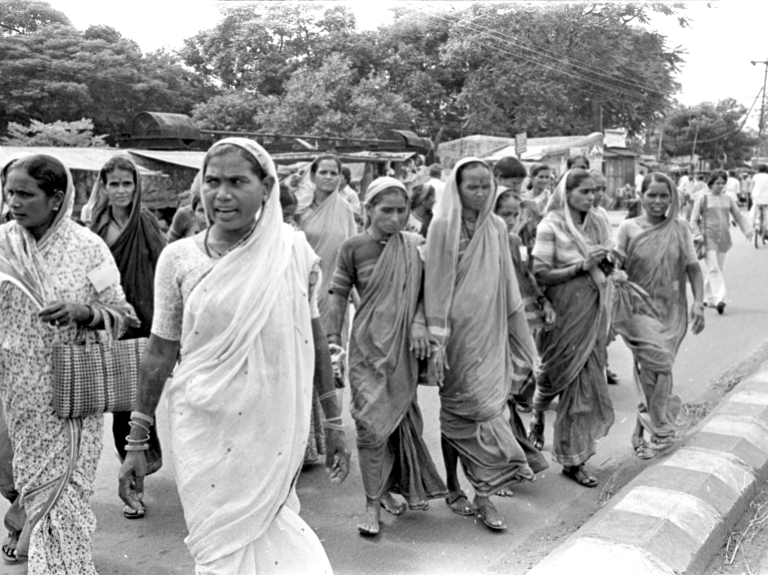 A wide shot shows a large group of women in India marching in protest. The image is black and white, and the women wear a variety of robes and veils.