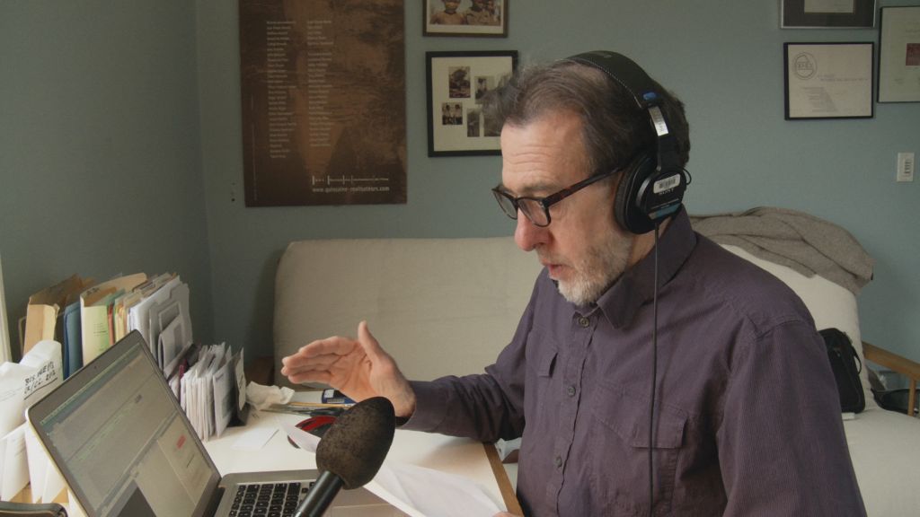 Filmmaker Ross McElwee sits at a laptop recording voiceover. He is a white man with grey hair and a white beard. He is wearing a brown collared short and headphones. There is a microphone on the desk beside the laptop.