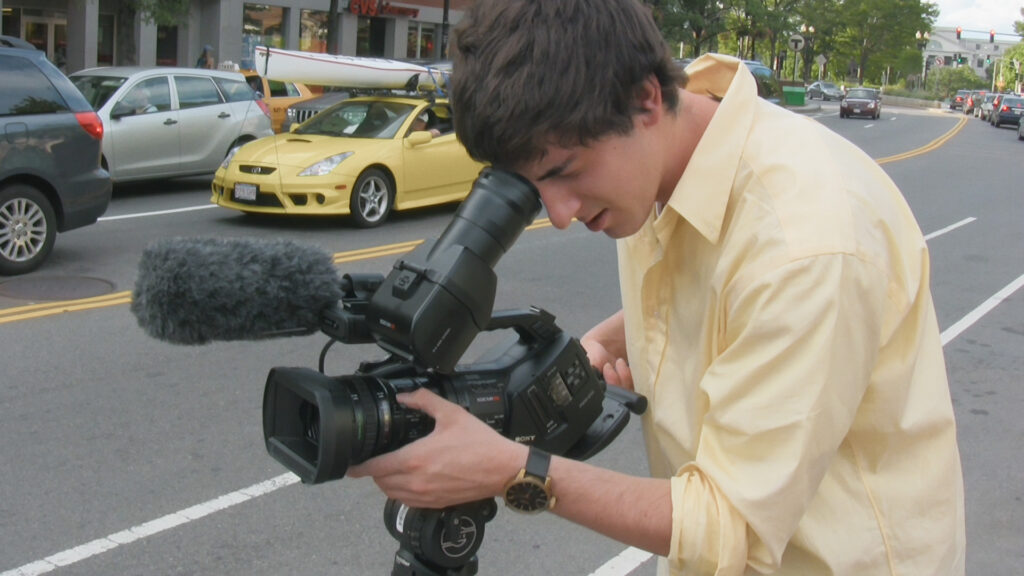 A photo of Ross McElwee's son Adrian as he records a scene on a camera. He is a young white man wearing a light yellow shirt and has brown hair. He is looking into the viewfinder and pointing the camera down a city street.
