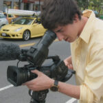 A photo of Ross McElwee's son Adrian as he records a scene on a camera. He is a young white man wearing a light yellow shirt and has brown hair. He is looking into the viewfinder and pointing the camera down a city street.