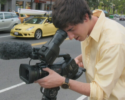A photo of Ross McElwee's son Adrian as he records a scene on a camera. He is a young white man wearing a light yellow shirt and has brown hair. He is looking into the viewfinder and pointing the camera down a city street.