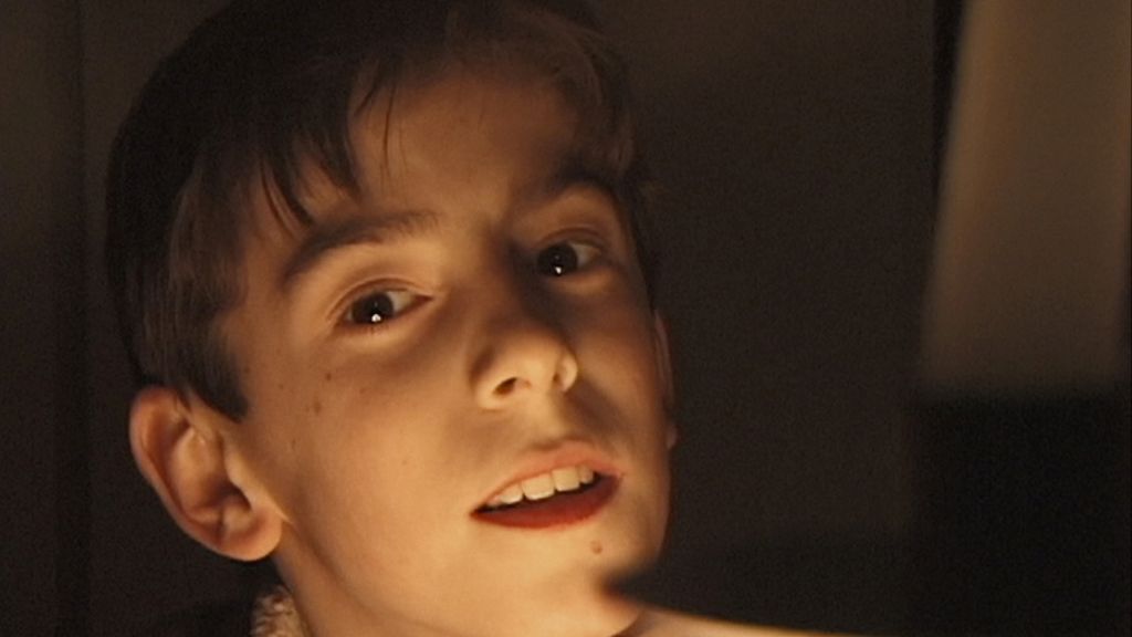 A close-up archival image from a home video shows young Adrian McElwee looking into the camera. He is a white boy with brown hair and brown eyes. The scene is dark but his face is bright and he seems happy.