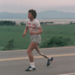 An archival image shows Terry Fox running along a rural street, wearing a white t-shirt and jogging shorts.
